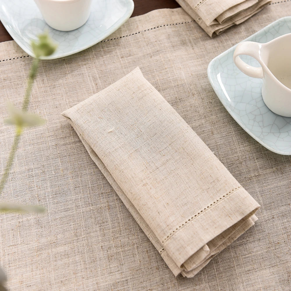 A stack of beige linen napkins resting on a neutral-toned tablecloth with a ceramic cup and plate nearby.