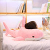 A pink giant shark plush toy lying on a white surface next to a person's hand holding a smartphone.