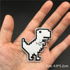 A hand holds a white embroidered punk badge shaped like a dinosaur.