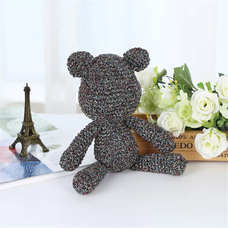 A small, dark purple textured teddy bear made from metallic silk crocheting and knitting yarn sitting on a white surface.