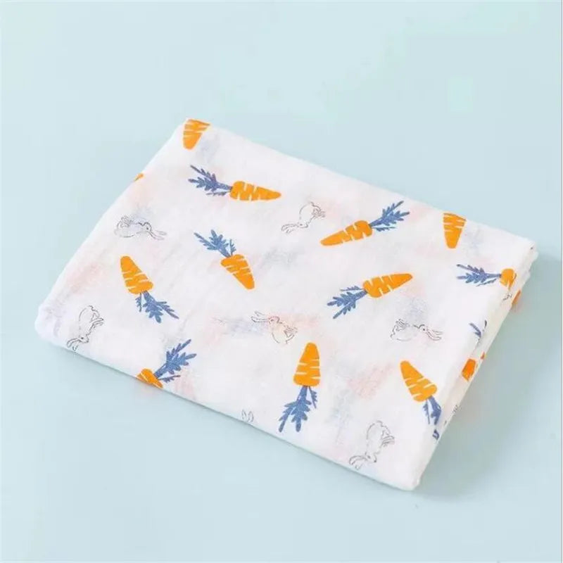 A folded white cotton muslin swaddle blanket with a pattern of orange and blue carrots.