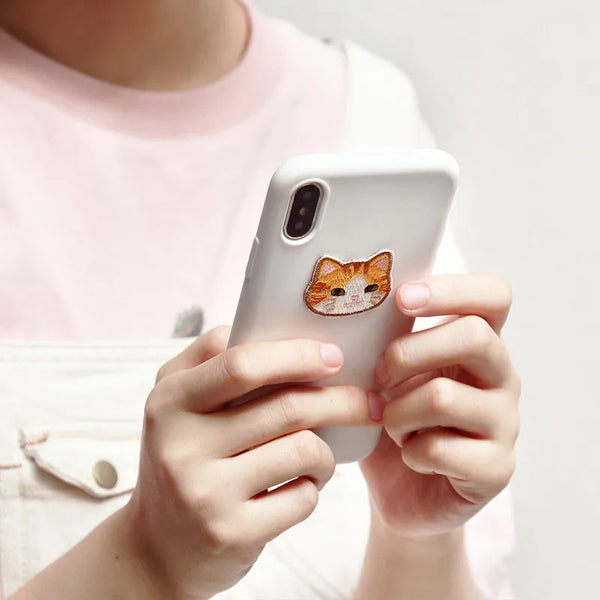 A person holds a white smartphone displaying an orange embroidered cat iron-on patch.