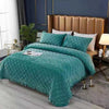 A teal quilted velvet bedspread is draped over a bed with a gold upholstered headboard and dark blue walls.
