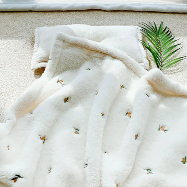 A white coral fleece flannel blanket with small orange and green floral embroidery is displayed on a light beige surface.
