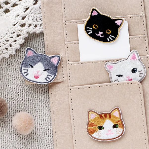 A collection of handmade embroidered cat iron-on patches in various colors and styles on a beige leather pouch.