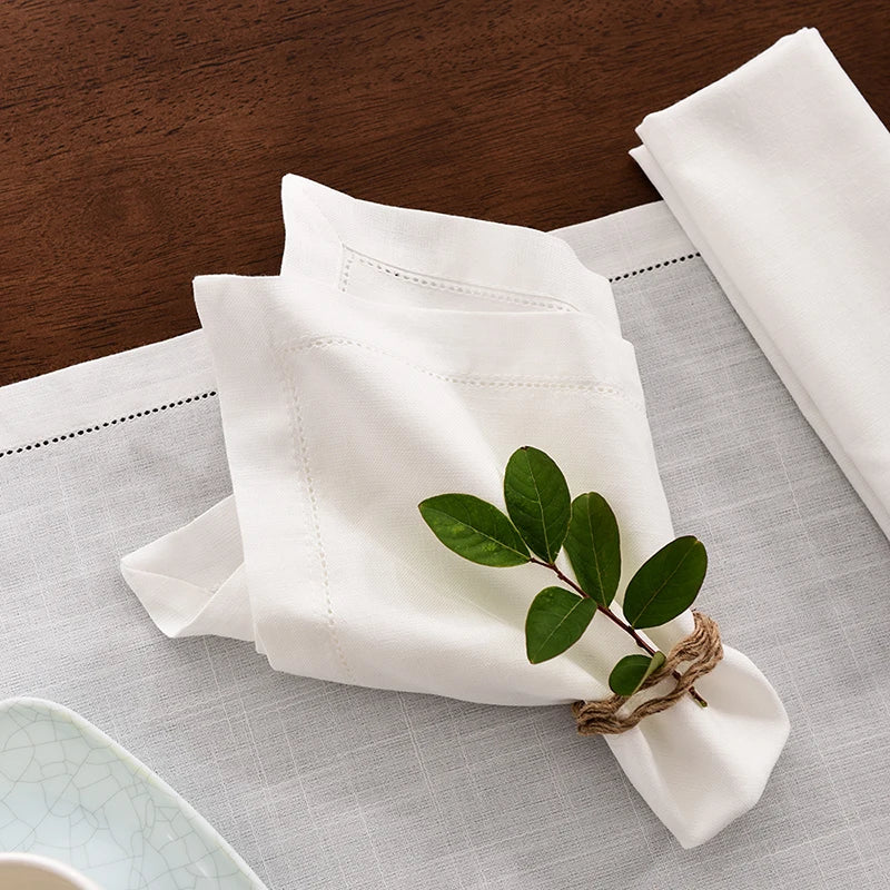 A stack of white linen napkins is displayed on a light gray cloth with a green leaf garnish.