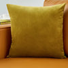 A square olive green velvet decorative cushion resting on an orange sofa.