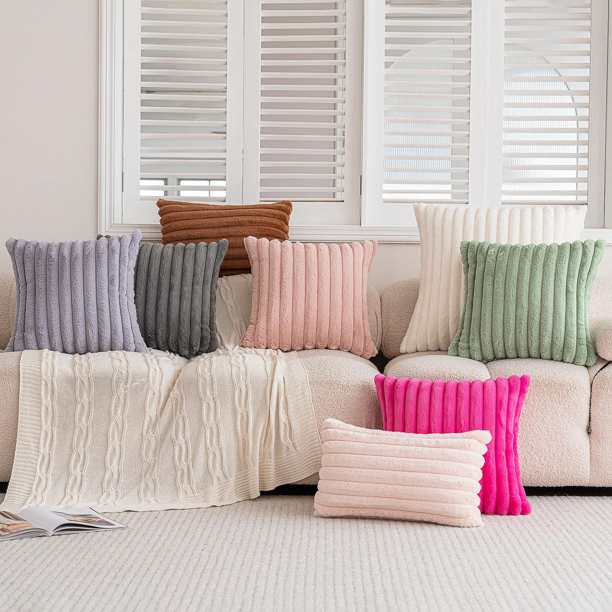 A collection of textured, vertical faux rabbit fur cushion covers in shades of gray, pink, and green displayed on a light-col