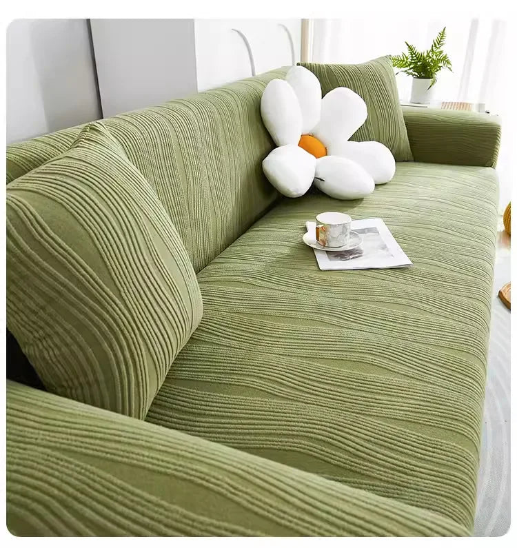 A textured olive green stretchable sofa cover with white decorative pillows and a cup on the seat.