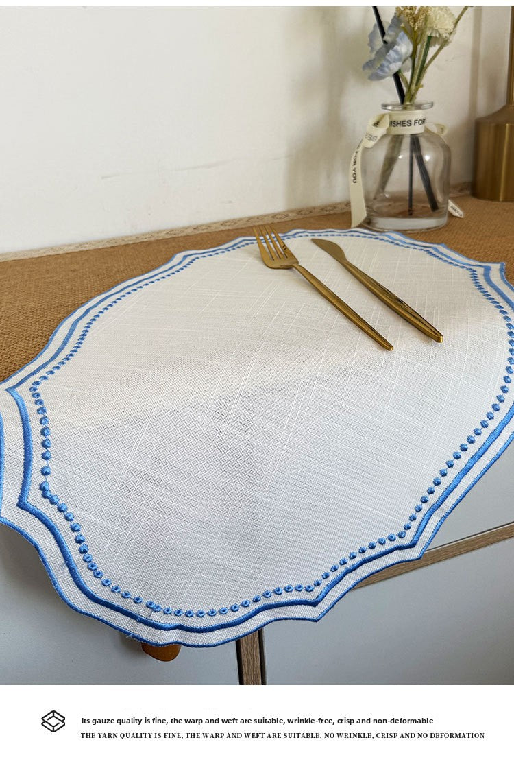 A white cotton linen placemat with blue embroidery and gold cutlery set on a wooden table.