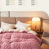 A pink quilted fleece blanket is displayed on a bed with white pillows and a bedside lamp.