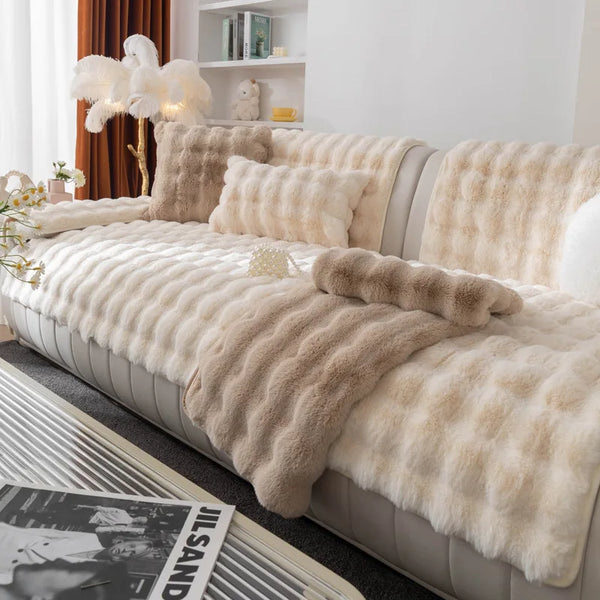 A beige plush rabbit fur sofa cushion cover draped over a light gray sofa with white pillows and a wooden lamp in a bright ro