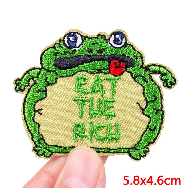 An embroidered patch featuring a green cartoon frog with a red tongue, with the text Eat The Rich stitched below.