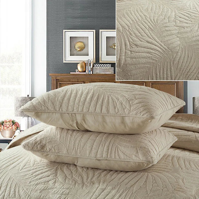 A set of beige embroidered cotton quilted pillow covers resting on a bed with a gray wall and wooden furniture.