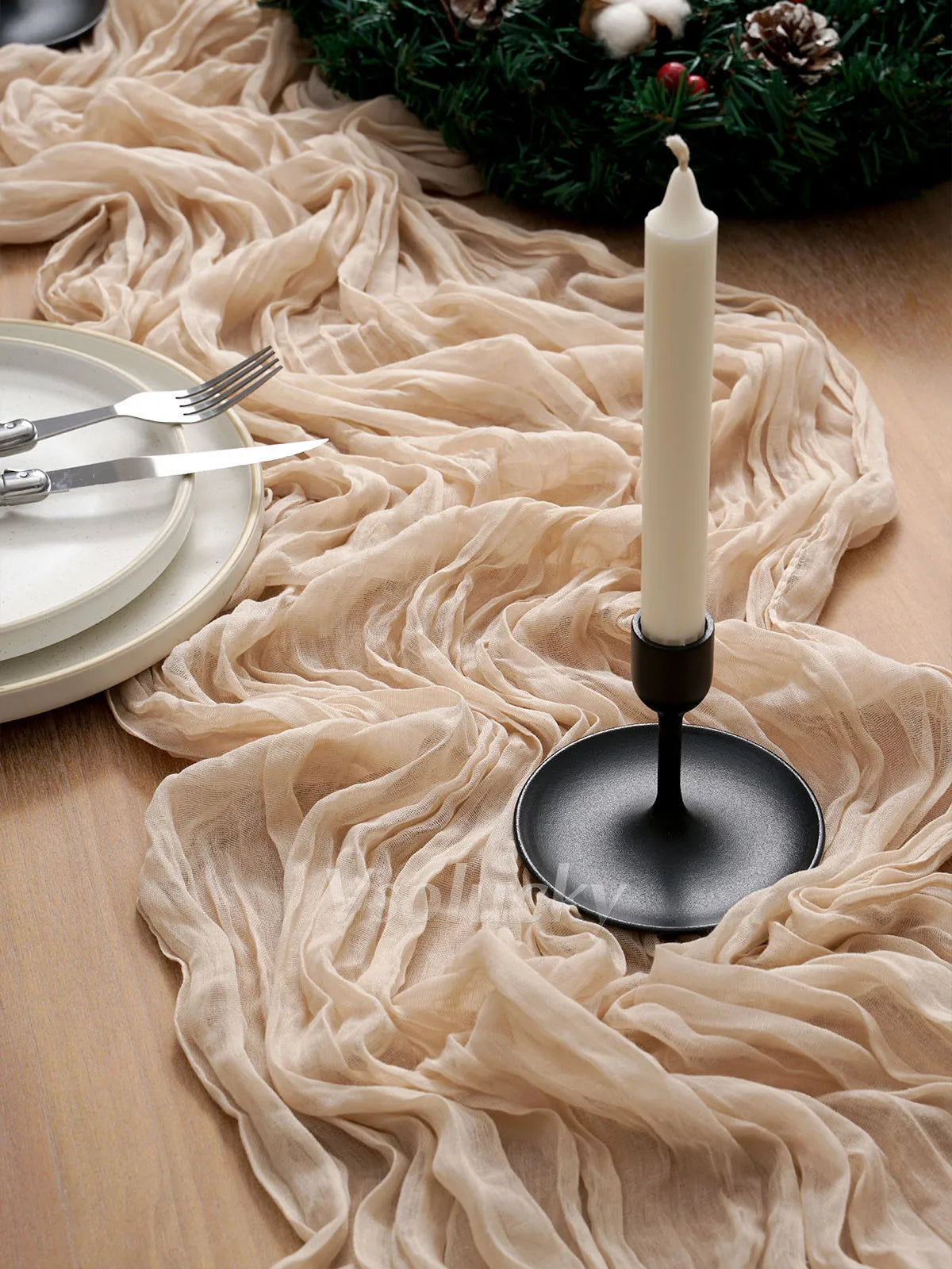 A beige crinkled cheesecloth table runner flows across a wooden surface next to a white candle and silverware.