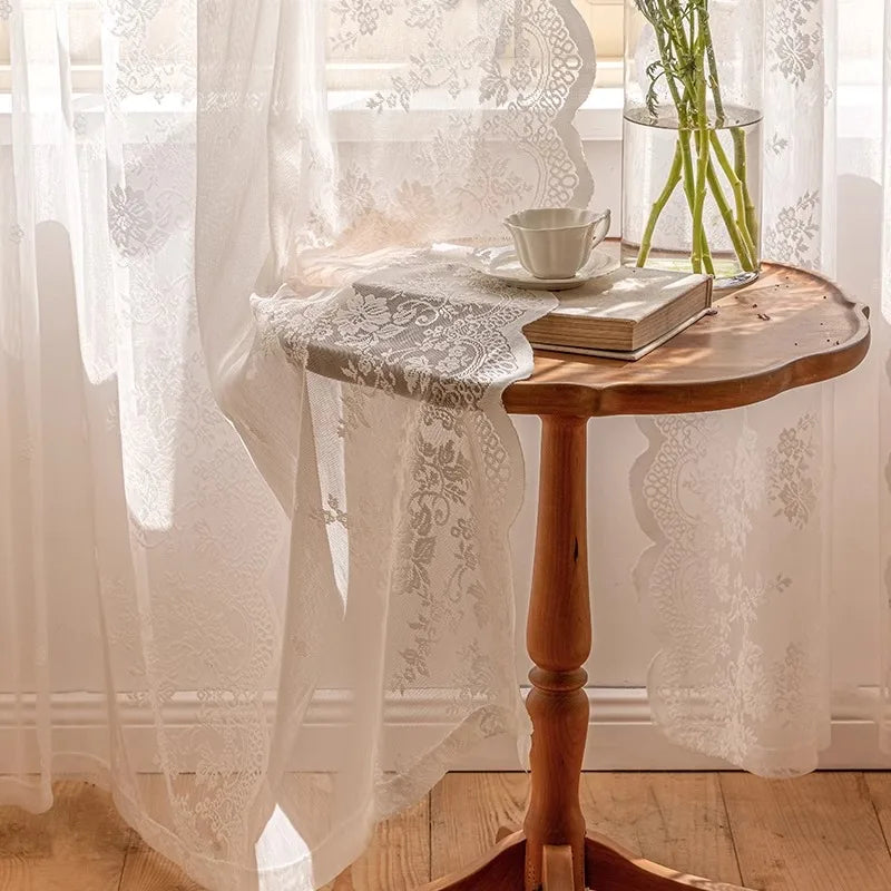 A wooden table with a glass vase and a stack of books is positioned in front of sheer floral lace curtains.