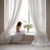 A young girl in a white dress looks out of a window draped with sheer white tulle curtains.