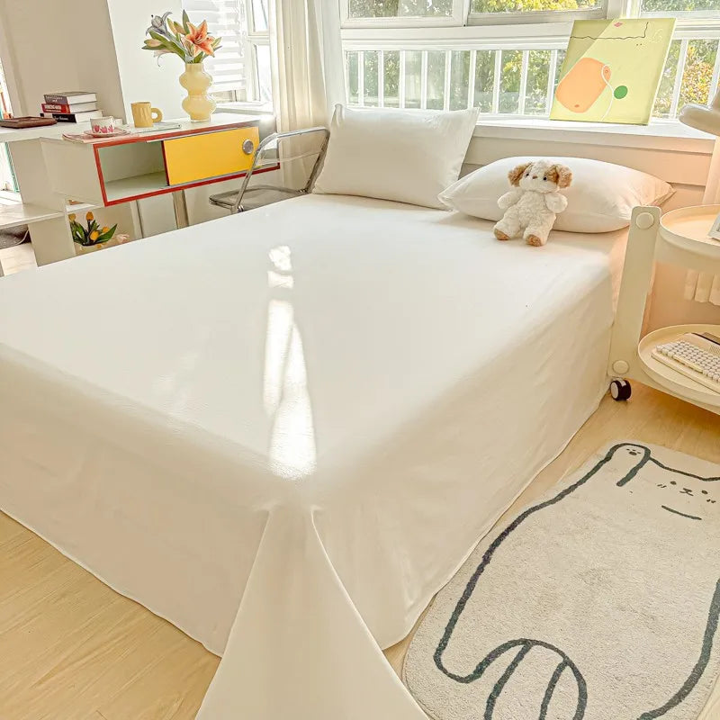A white organic cotton bedding set with a flat sheet and pillowcases is displayed on a bed in a bright room.