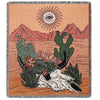 A woven throw blanket featuring a desert landscape with mountains, a sun motif, and various cacti and succulents.