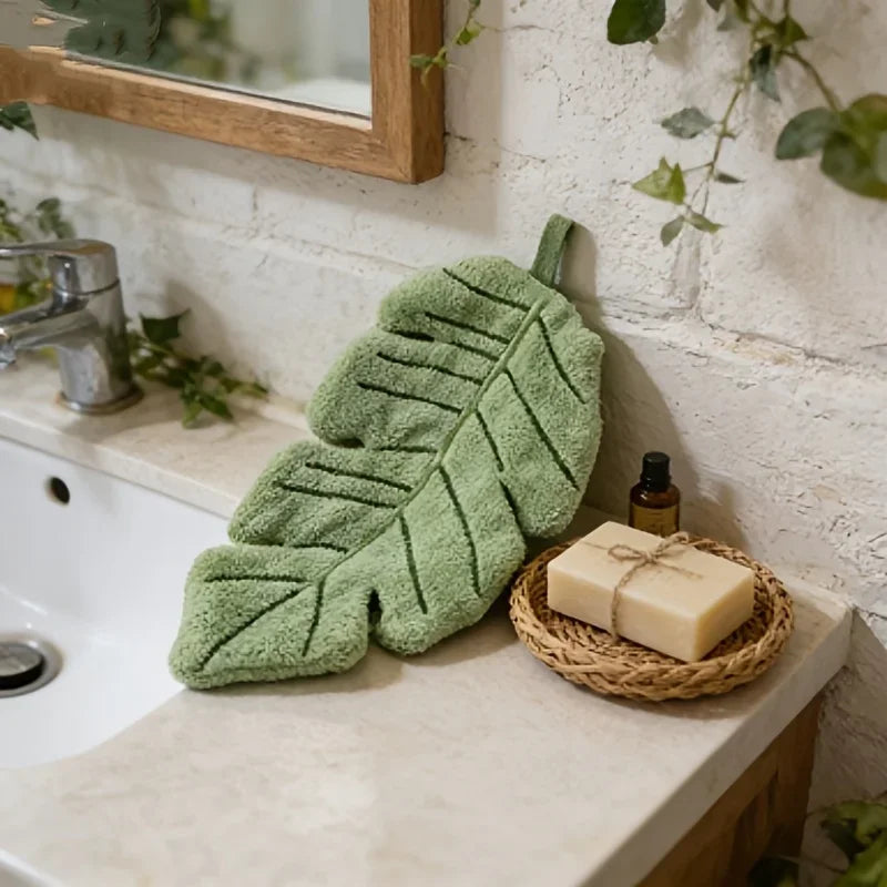 A green decorative banana leaf hanging hand towel next to a bar of soap on a bathroom counter.