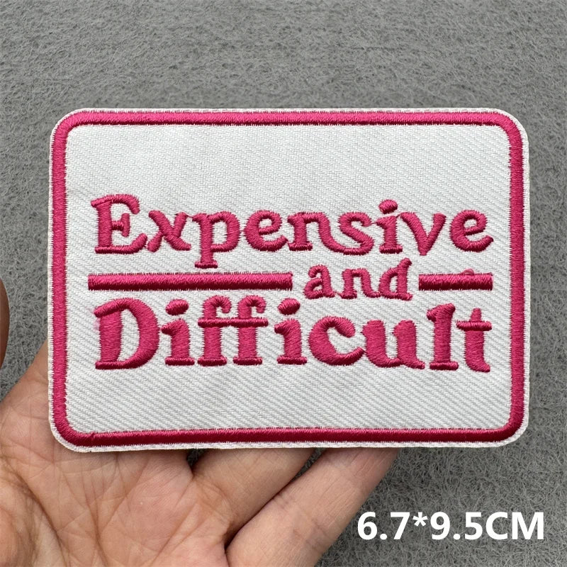 Pink embroidered patch with the text Expensive and Difficult on a white fabric background