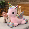 A pink unicorn plush toy with a rainbow horn sitting on a table next to a woman.