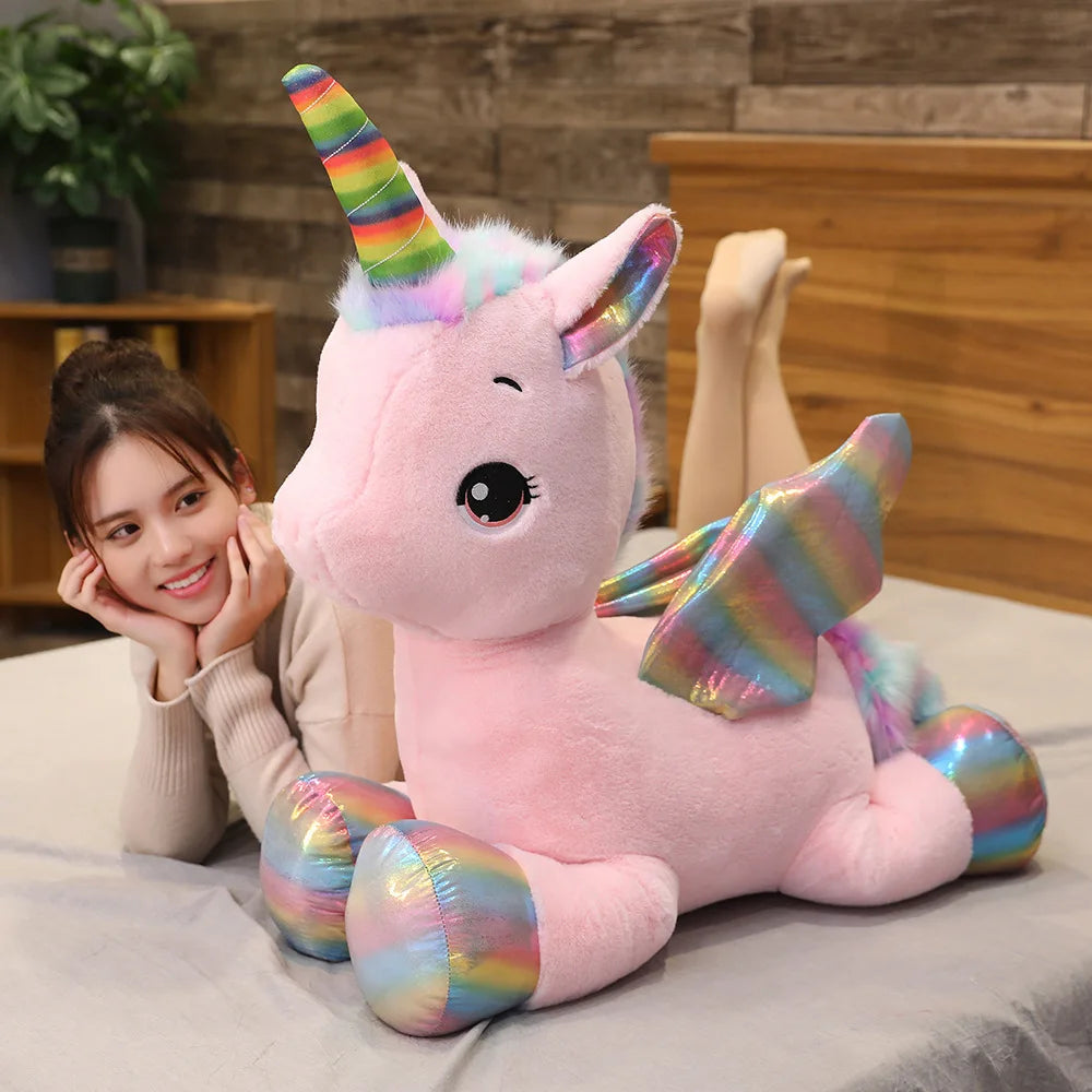 A pink unicorn plush toy with a rainbow horn sitting on a table next to a woman.