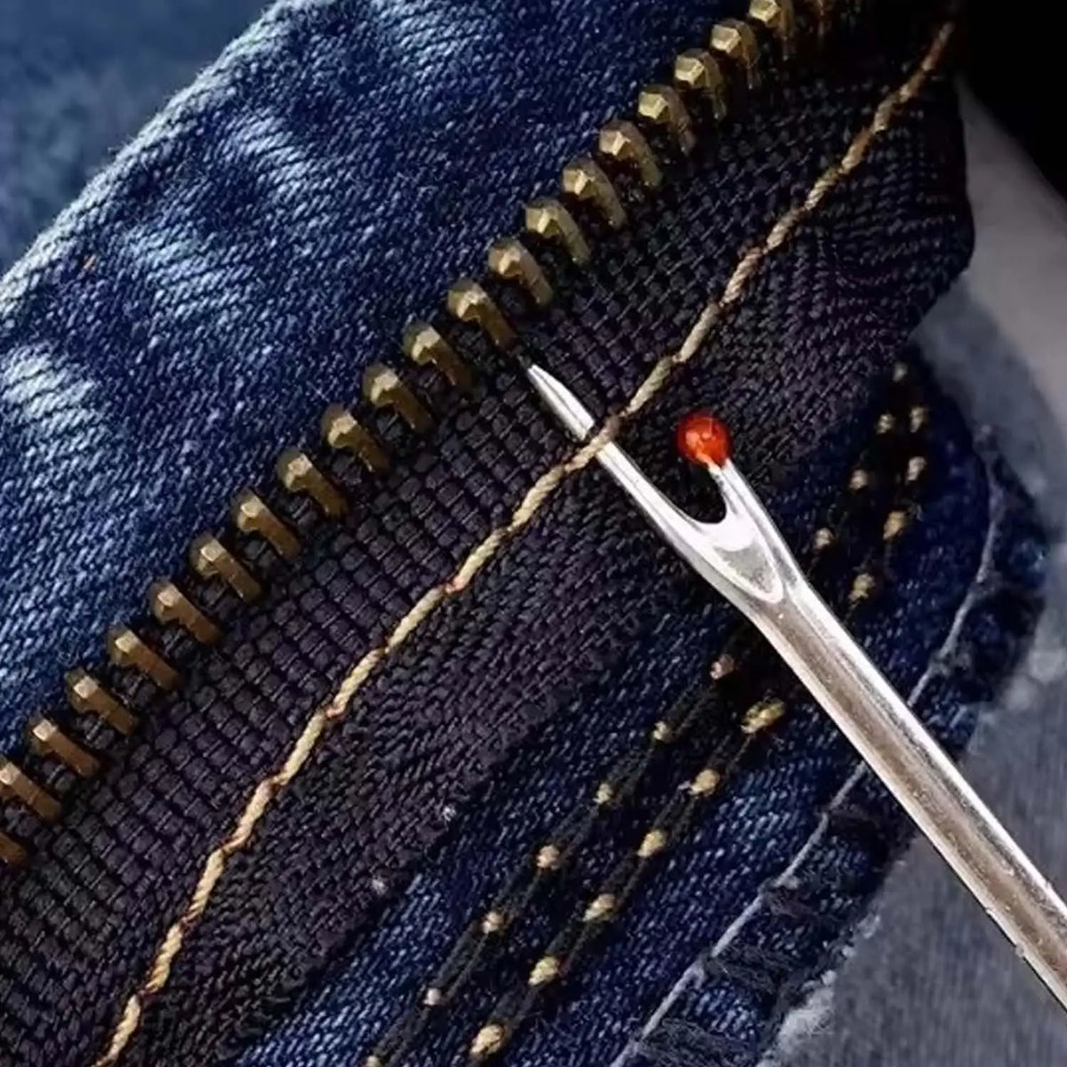 A close-up of dark denim fabric showing a stitched seam with a gold thread and a silver sewing needle.