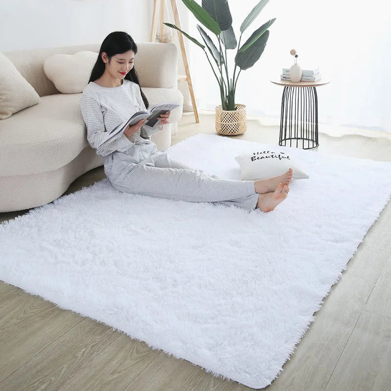 A woman reading a book on a white fluffy rug in a bright room with a large green plant.