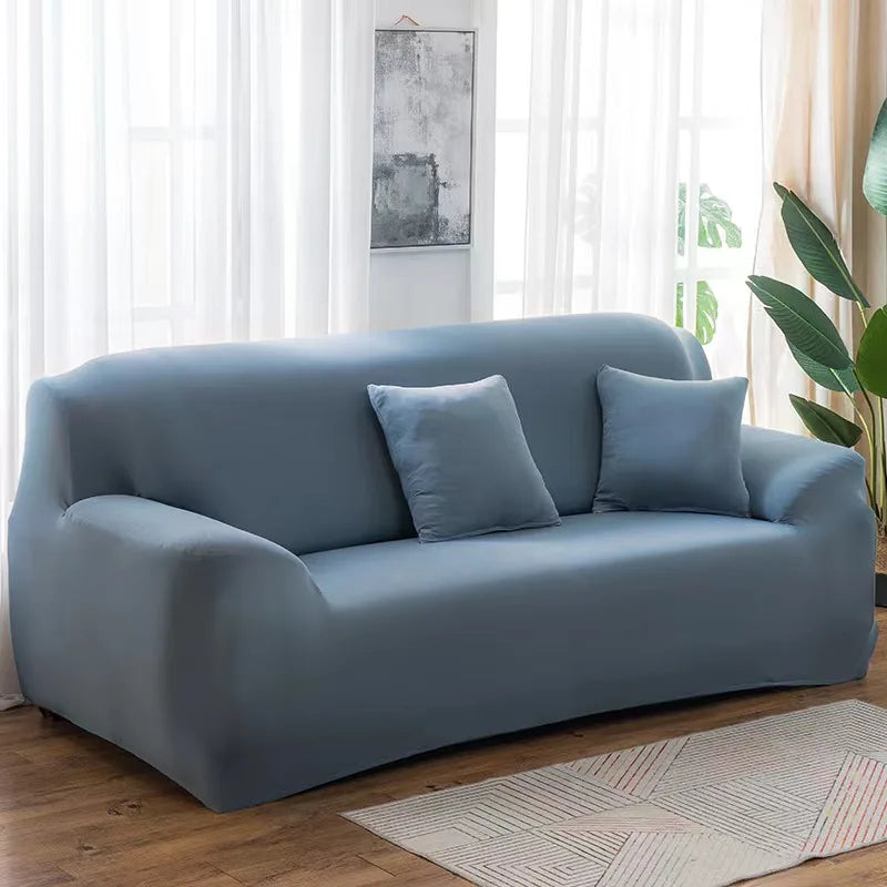 A modern, light blue stretch sofa cover with gray throw pillows placed in a bright living room.
