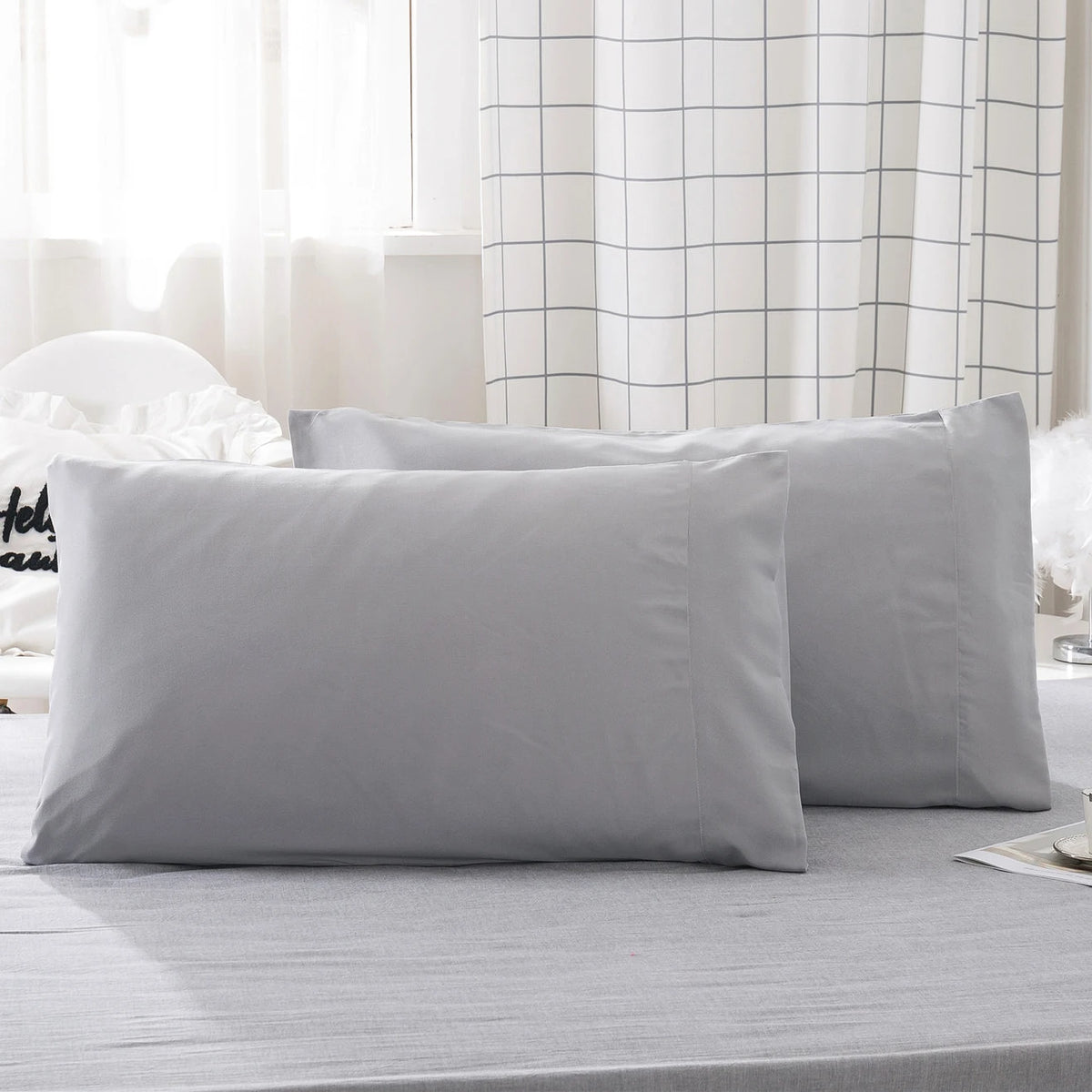 A stack of solid gray microfiber pillow covers on a bed with white curtains.