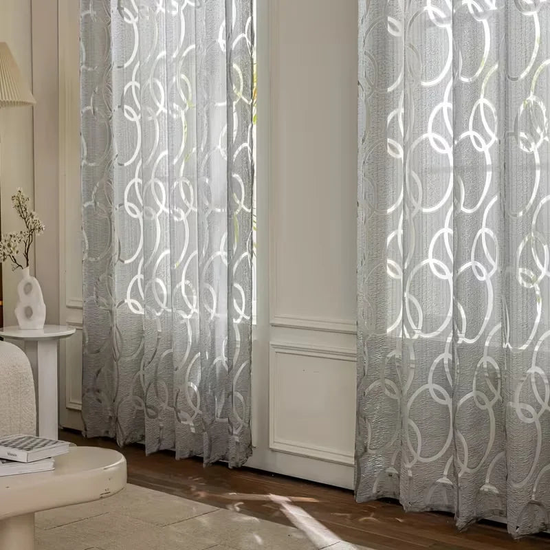 A pair of modern geometric embroidered sheer curtains in light gray hanging in a window.