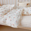 A white muslin blanket with a floral print is folded on a light pink surface next to a cream-colored storage basket.