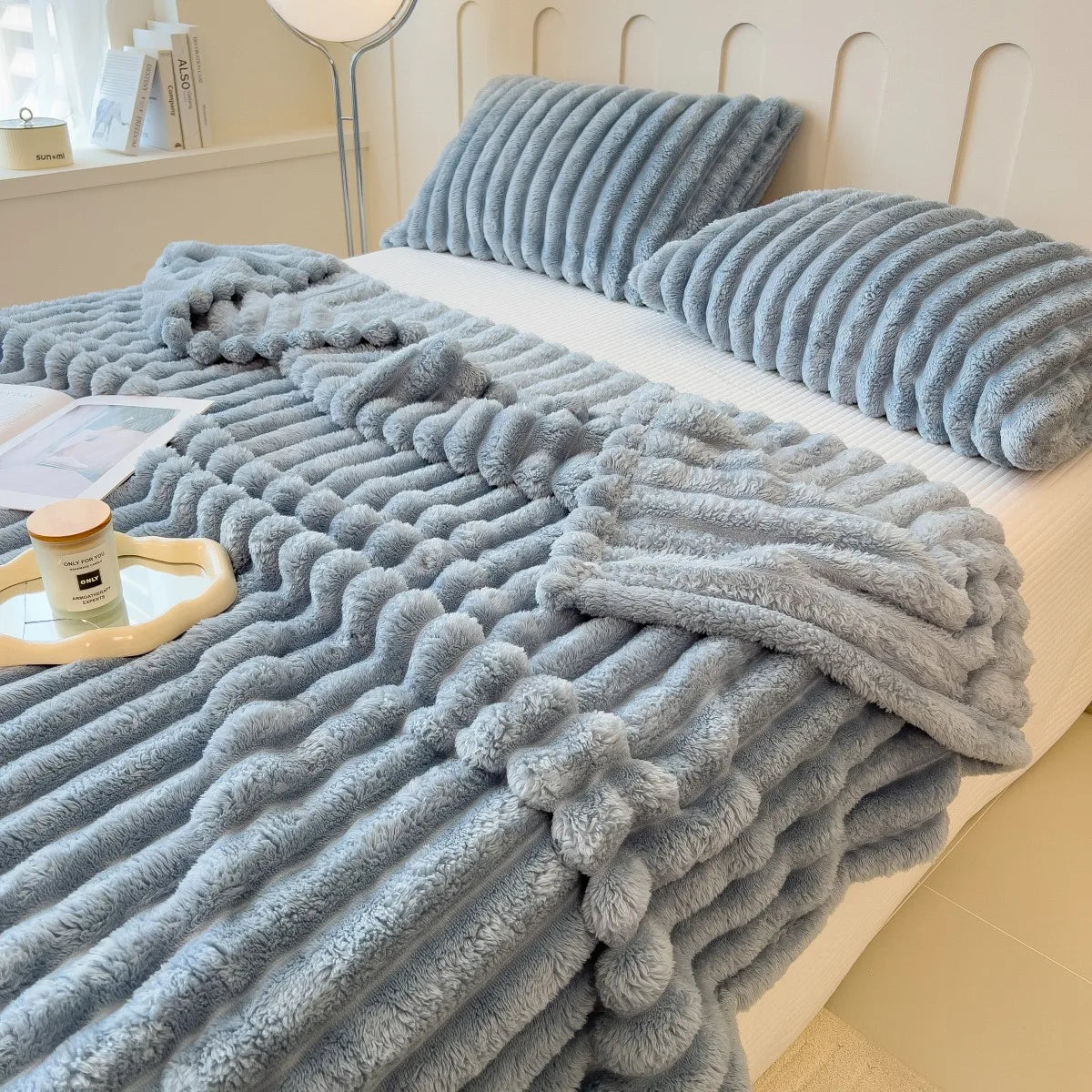 A thick, blue fuzzy blanket is draped over a white bed with matching striped pillows.