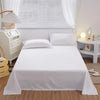 A white flat bed sheet is spread on a bed with white pillows, next to a window and bedside table.