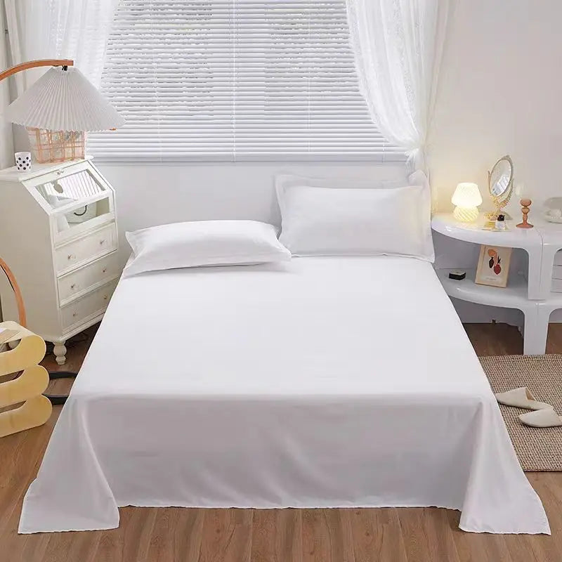 A white flat bed sheet is spread on a bed with white pillows, next to a window and bedside table.