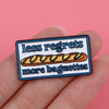 An enamel pin featuring the text less regrets more baguettes on a rectangular background.