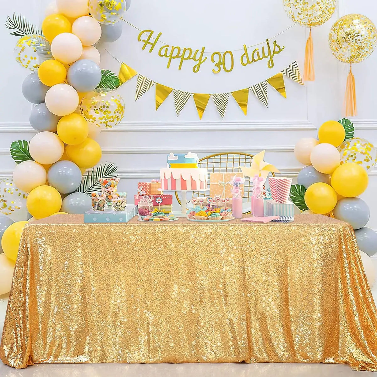A gold sequin rectangle table cover is displayed on a table, decorated with balloons and a banner reading Happy 30 days.