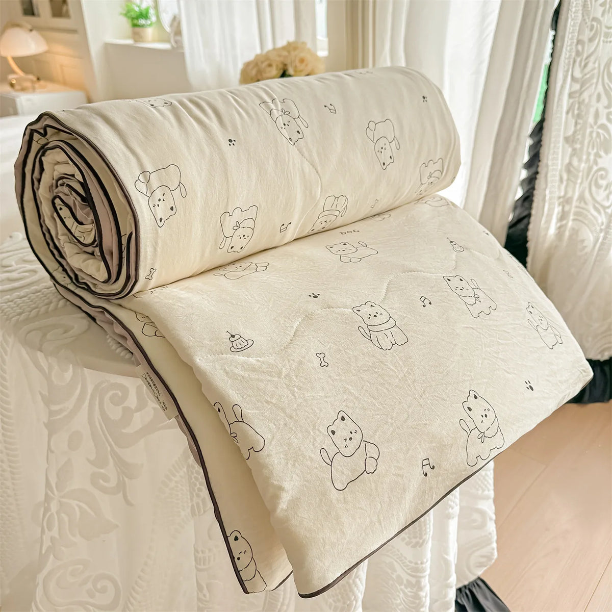 A rolled cream-colored quilt blanket with a pastoral print featuring small animal figures is displayed on a white surface.