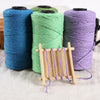 Three spools of cotton blend yarn in teal, green, and purple next to a wooden loom.
