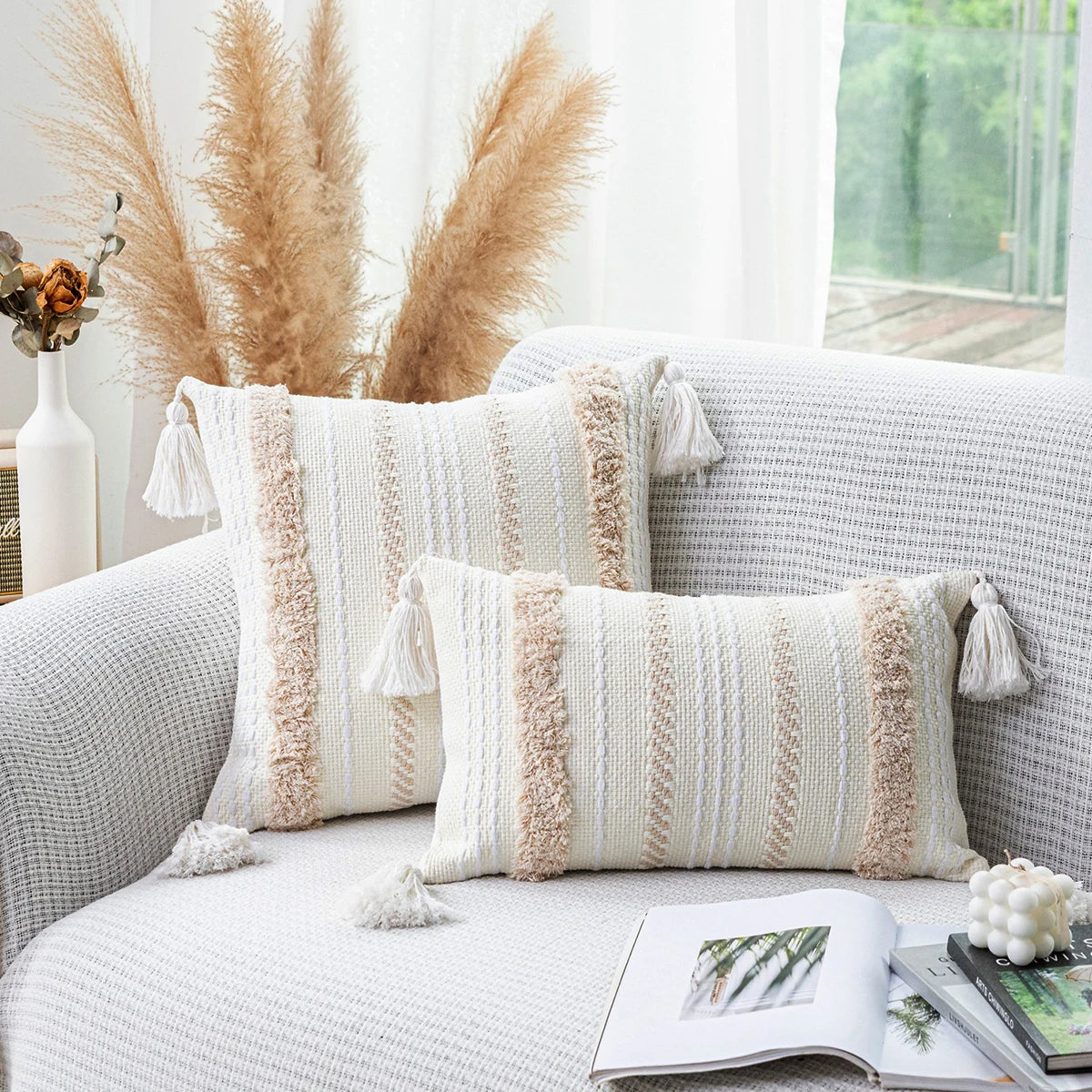 A beige tufted throw pillow cover with fringed tassels resting on a light gray sofa.