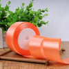 A roll of bright orange satin craft ribbon resting on a wooden surface with green foliage in the background.