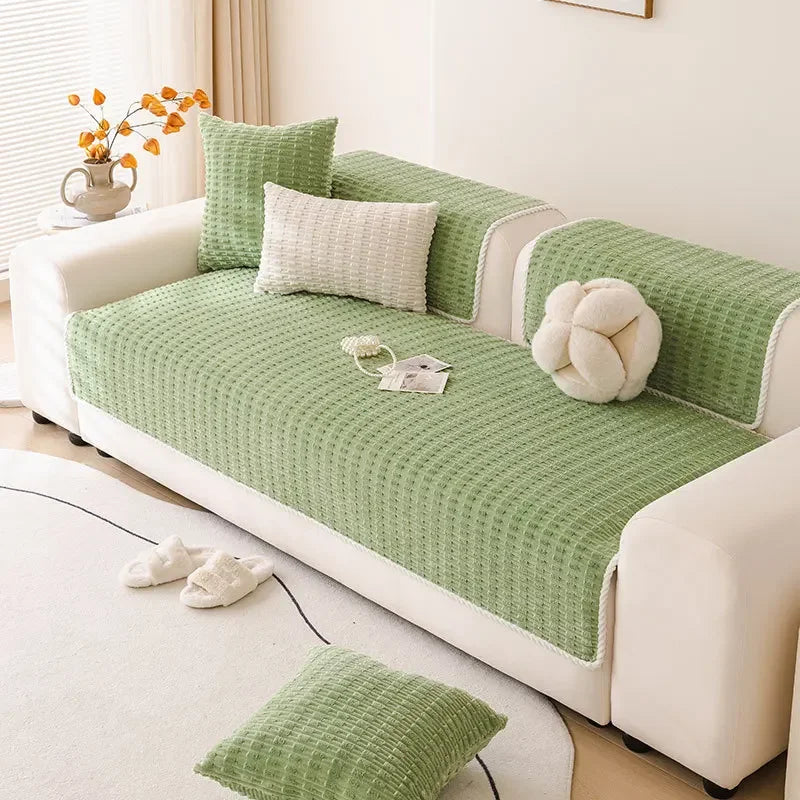 A light green quilted non-slip sofa cover with a white cushion and decorative pillows on a white sofa.