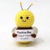 A yellow crocheted bee plush toy with black antennae and a sign that reads Positive Bee I bee-believe in you