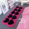 A pink fuzzy heart-shaped floor mat is displayed on a gray carpet next to a pink bed and framed art.