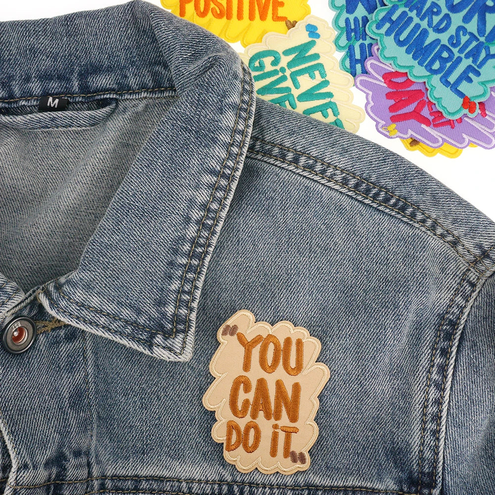 A close-up of a denim jacket featuring an iron-on embroidered patch that reads You Can Do It