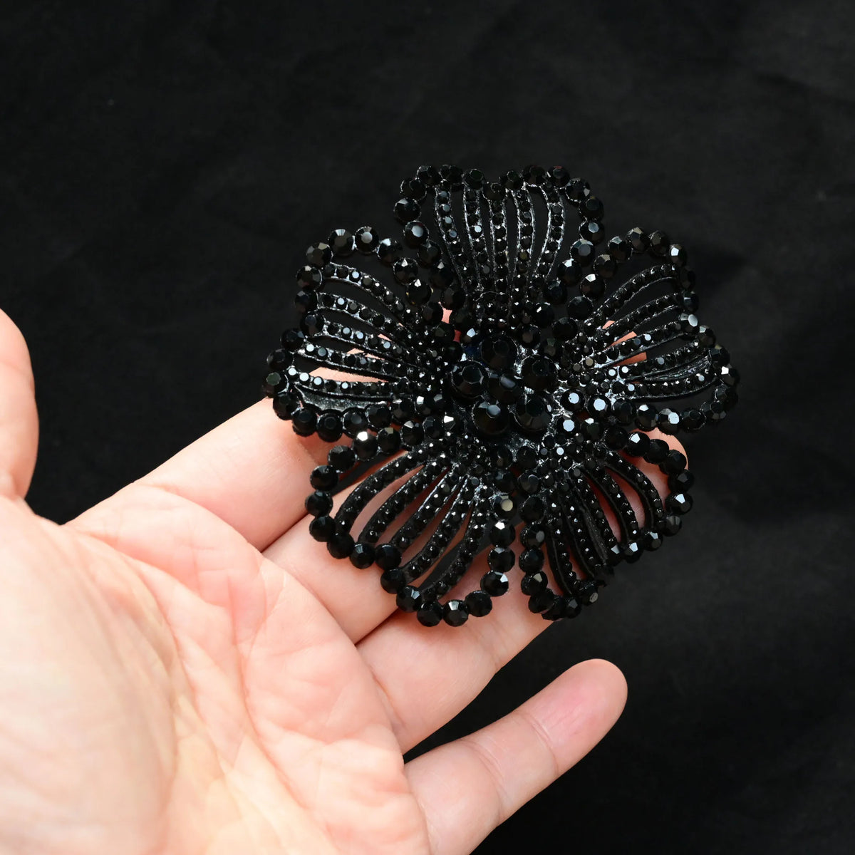 A hand holds a black rhinestone flower applique patch with intricate detailing.