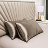 A set of smooth satin silk pillow covers in a muted champagne color resting on a bed with a lamp nearby.