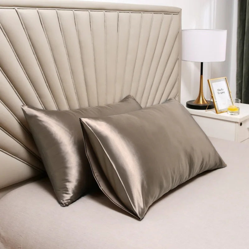 A set of smooth satin silk pillow covers in a muted champagne color resting on a bed with a lamp nearby.