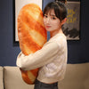 A young woman is holding a large, orange French bread plush pillow against her chest indoors.
